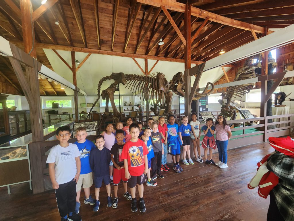 In front is a group of second grade students in a large wooden building. Behind them are the bones of a dinosaur.