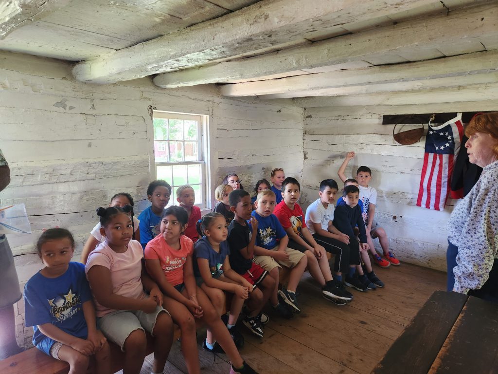 A group of second grade students sit on a wooden bench in a small room and listen as the woman in front of the room talks.