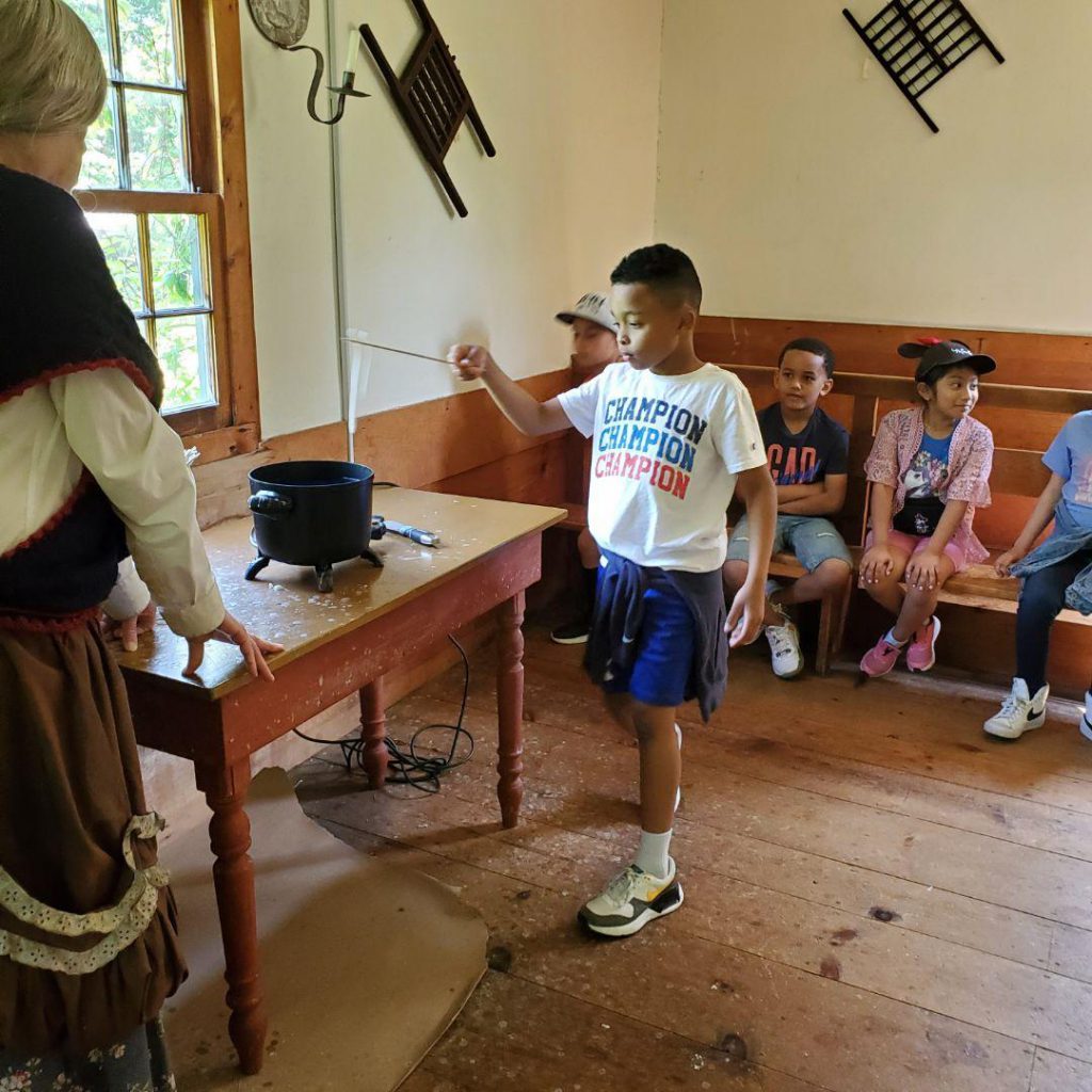 A boy with blue shorts and white tshirt stands in front of the group of students with a stick. He is dipping it into wax to create a candle.