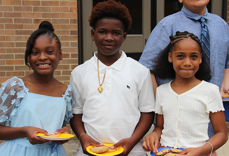 Three fifth-grade students stand together smiling, each holding cookies on a plate.