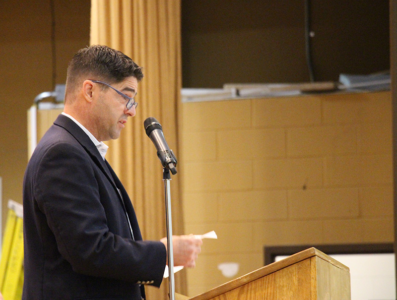 A man in a dark suit jacket, with dark hair and glasses, speaks at a podium.