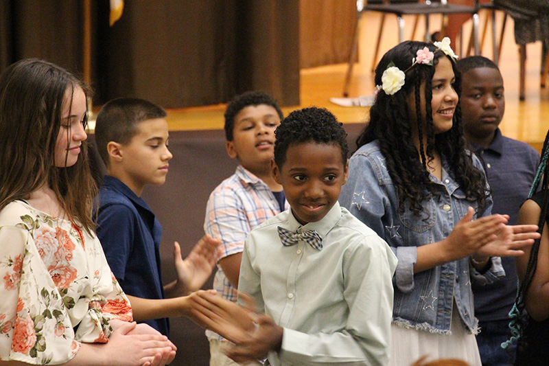 A group of fifth-grade students stand and clap. They are smiling.