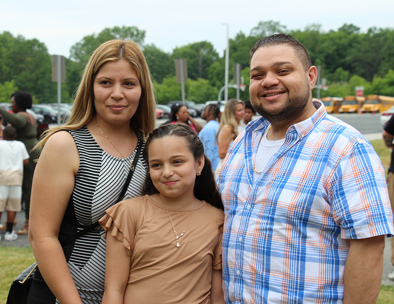 A man and woman flank a fifth-grade girl. They are standing outside and all are smiling.