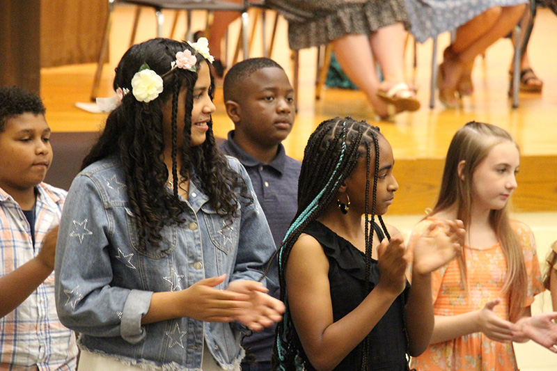 A group of fifth-grade students stand and clap. One girl has a ring of flowers in her hair.