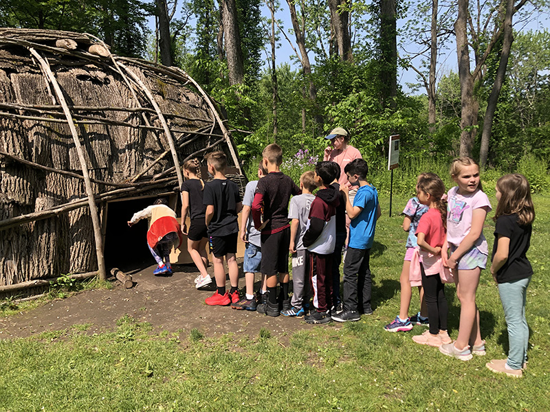 A line of third-grade students stand waiting to go into a hut. The girl in front is bending over to get into it. 