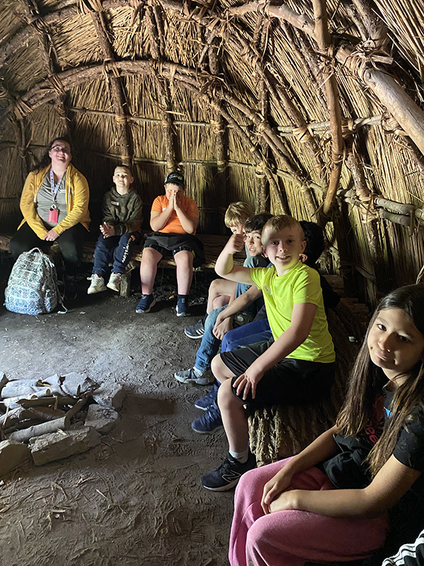 Students and adults sit inside a hut.