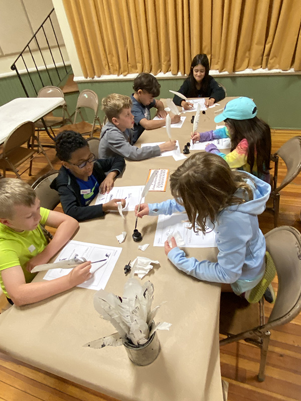 A group of third grade students write with quills and ink.