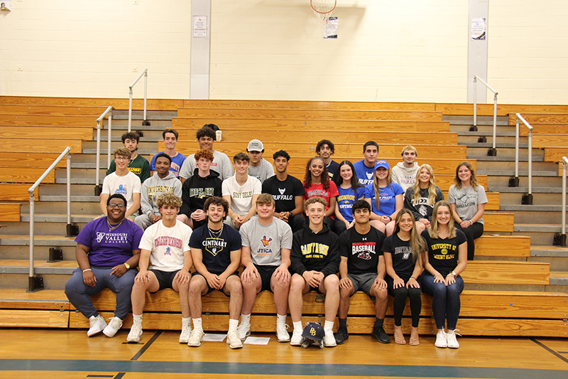 A group of 25 high school students, all wearing athletic shirts, sit on bleachers in three rows.