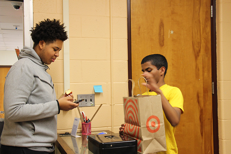 A young man uses his phone to pay for an item in the store. Another young man on the right hands him a bag.