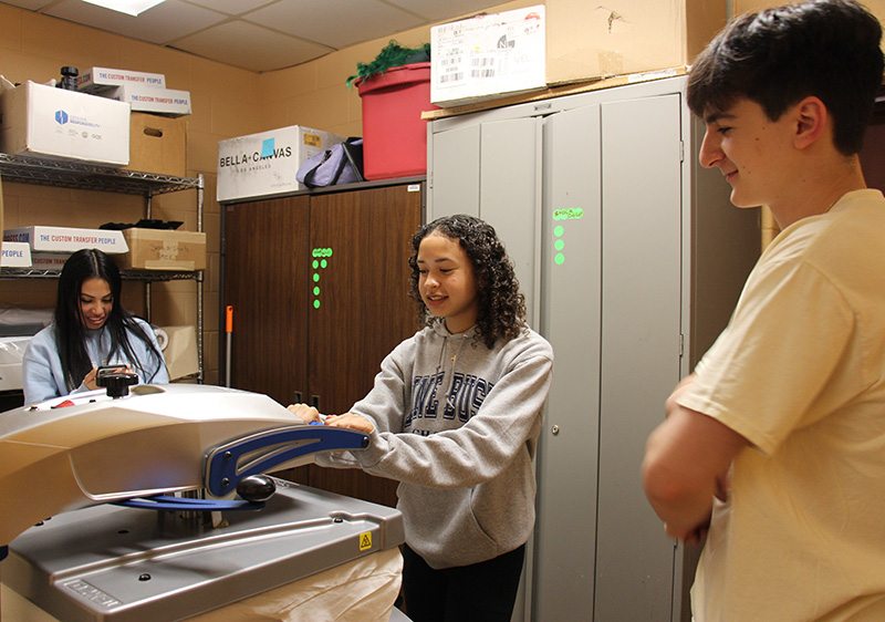 Three high school students stand in a room where the person in the center is using a heat press on a shirt.