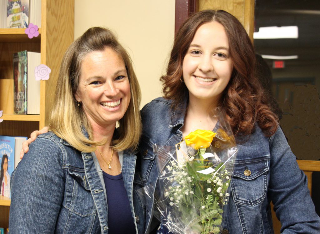 A woman with shoulder-length blonde hair, wearing a denim jacket smiles. A high school senior with long reddish hair, wearing a denim jacket. She is holding yellow flowers. Both are smiling and have their arms around each other.