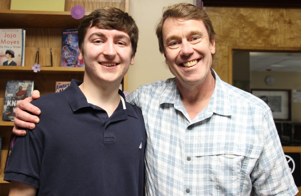 Two men stand together with the man on the right with his arm around the young man on the left. The high school senior on the left has a blue polo shirt on. He has short brown hair. The man on the right has a plaid shirt on, has short sandy hair. Both are smiling.