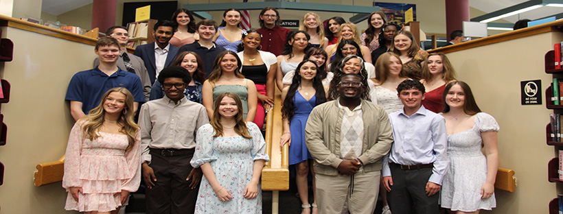 A group of 27 high school seniors, all dressed up, stand on the stairs in a library.
