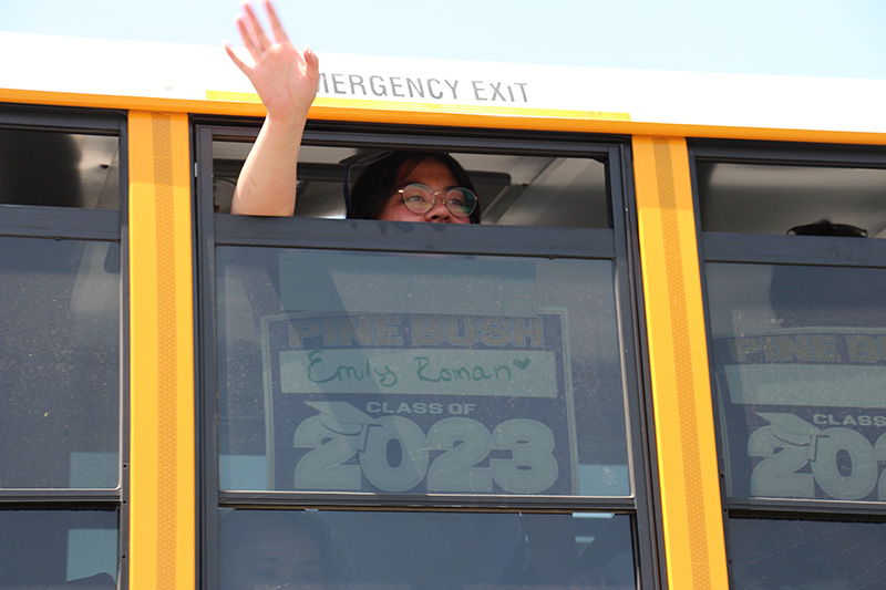 A high school senior holds a sign in a bus that says Senior 2023. She waves out the window of the yellow school bus.