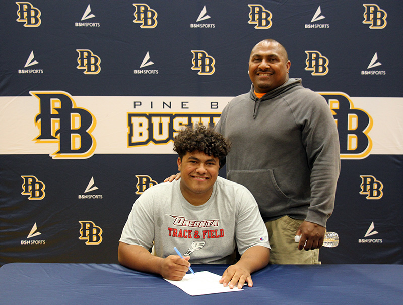 A high school young man sits at a table signing a piece of paper. He is smiling. Next to him is a man smiling and the backdrop says Pine Bush Bushmen.