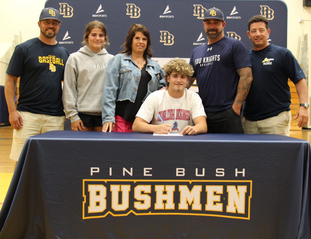 A high school senior sits at a desk that has a tablecloth that says Pine Bush Bushmen. He is surrounded by five adults standing behind him.