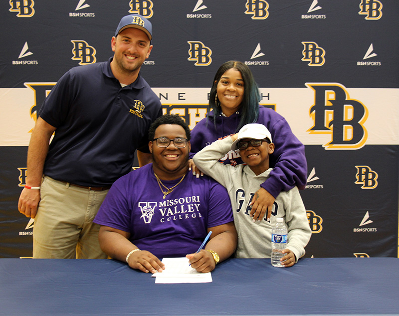 A high school young man smiles broadly as he sits at a table signing a paper. Standing next to him are a man with his arm around his shoulder, a woman and a younger boy. All are smiling.