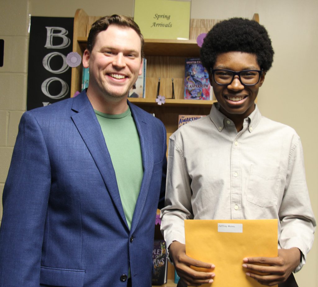Two men stand together. One is wearing a green shirt and blue jacket. The young man on the right has glasses and is wearing a white button-down shirt. He is holding a manilla envelope. Both are smiling.