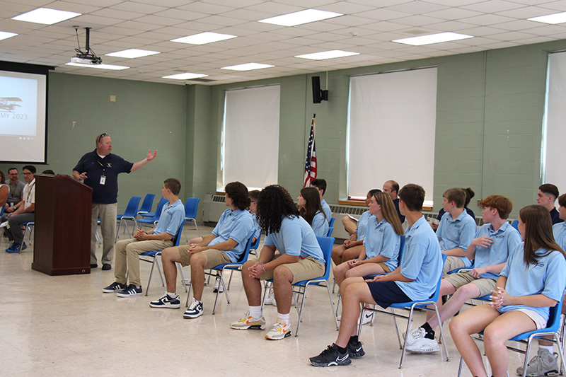 A man at the far left, wearing tan pants and a navy blue shirt, gestures to the high school students, all wearing light blue shirts and sitting listening to him.