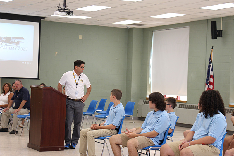 A man in a white polo shirt stands at a podium and is turned to talk to a group of high school students sitting in chairs. They are all wearing light blue shirts.