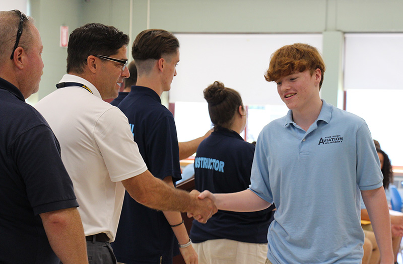 A high school young man with short red hair, shakes hands with a man wearing a white polo shirt. They are both smiling.