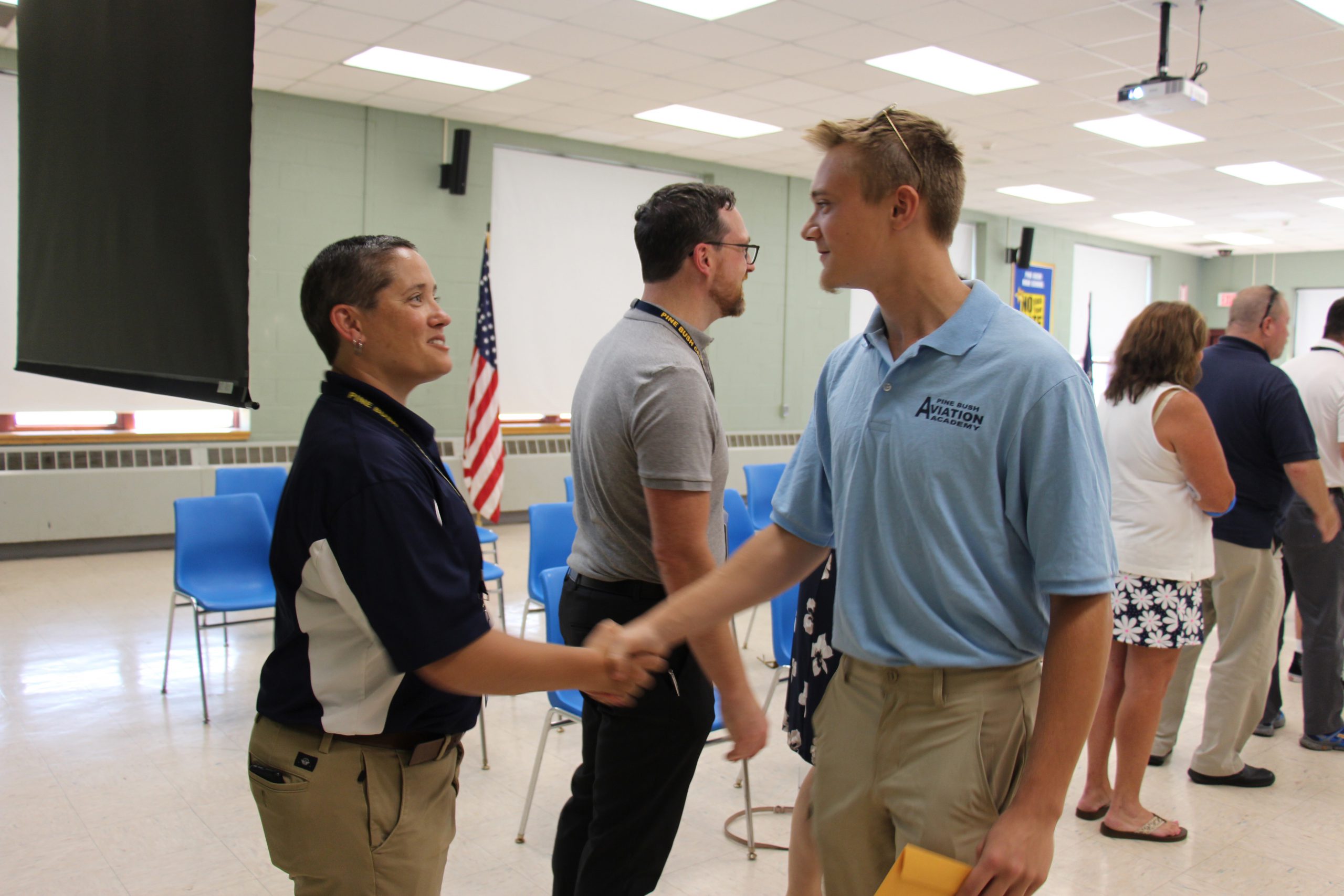 A tall high school young man wearing a light blue shirt and tan pants, shakes hands with a woman with short dark hair and wearing a blue and white shirt.