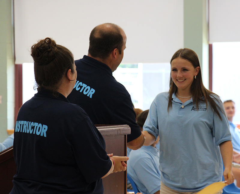 A high school young woman with long hair, wearing a light blue shirt, shakes hands with a man wearing a dark blue shirt. She is smiling.