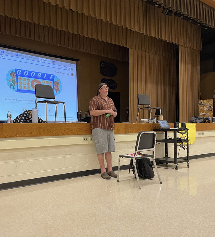 A man stands in front of a room of middle school students, speaking to them. Behind him is a screen.