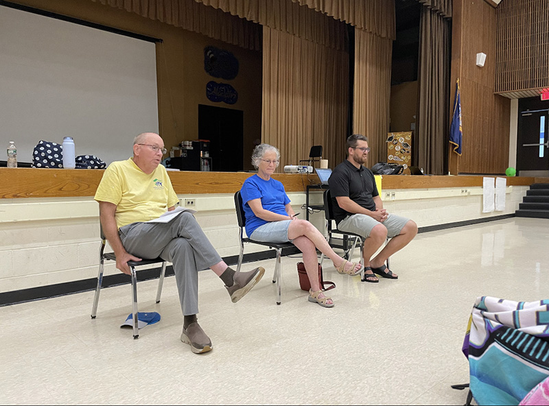 Three people sit in chairs in the front of an auditorium, speaking to a group of middle school kids.