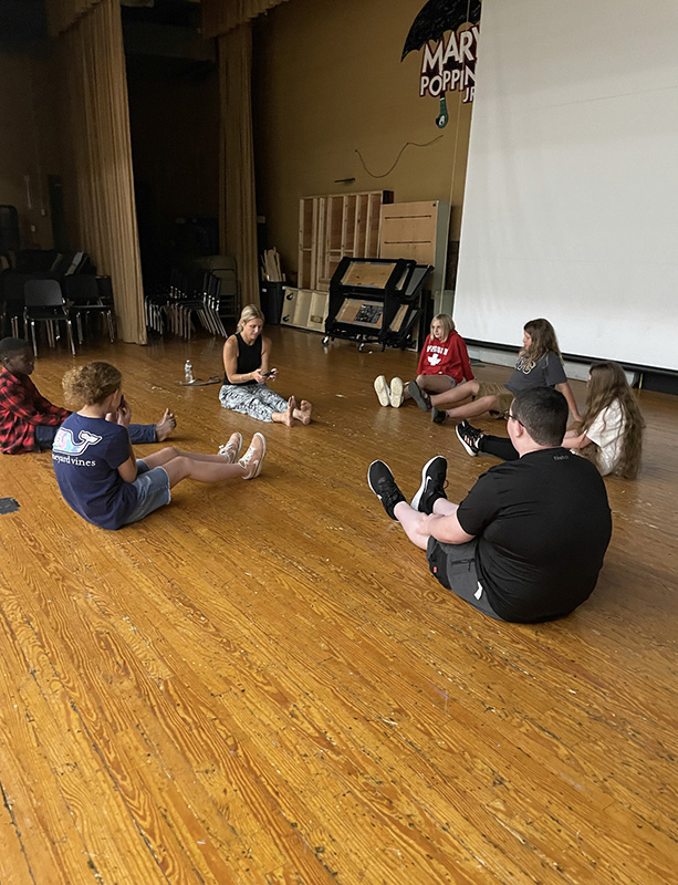 A group of six middle school students sit on the floor in a circle with a woman who is doing yoga with them.