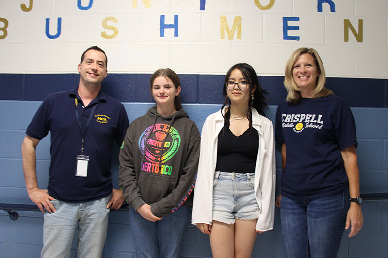 Two adults on either end smile broadly, a man at the left in a navy blue shirt with PB on the chest and a woman on the right with chin-length blonde hair, wearing a Crispell tshirt, with two middle school students in the center. The girl second from left has her hair pulled back in a ponytail and is wearing a gray sweatshirt with different colors in the center and the girl second from right has dark hair, glasses and is wearing shorts, a black shirt and white sweater.