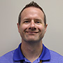 A man in a blue collared shirt smiling. He has short brown hair.