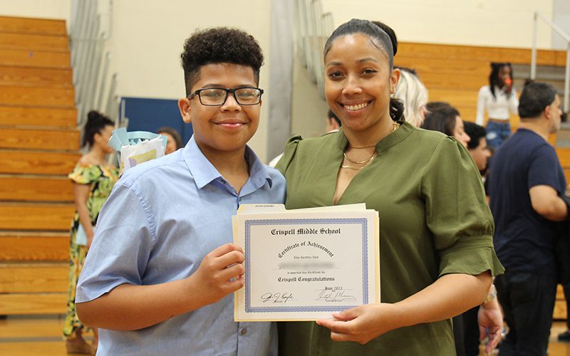 A young man with glasses wearing a blue button down shirt holds a certificate from the left while a woman on the right holds the other side of the certificate. She has her hair pulled back and is wearing a green dress. Both are smiling.
