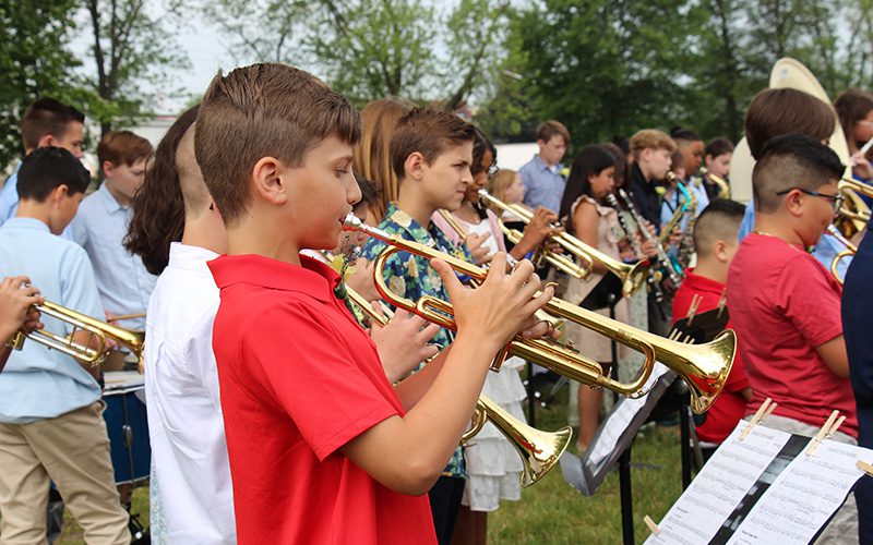 A group of fifth-grade musicians stand playing. The closest one is playing a trumpet. He is wearing a red shirt.
