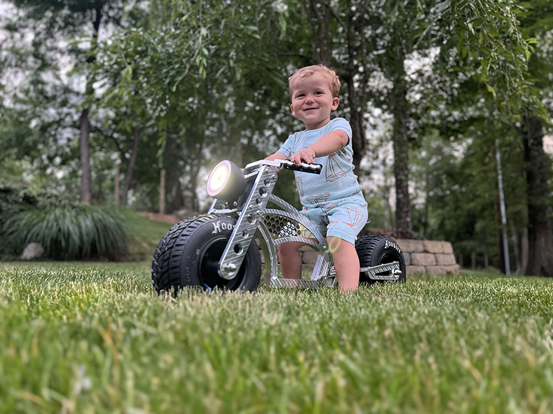 A toddler boy wearing blue shorts and shirt, smiles as he sits on a metal bike with large rubber wheels.