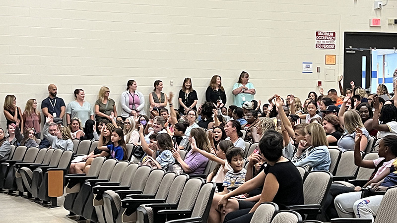 An auditorium with many adults and middle schools students sitting and listening. Several of the kids are raising their hands.