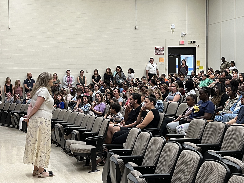 An auditorium filled with adults and middle school students listening as a woman in the front talks to them.