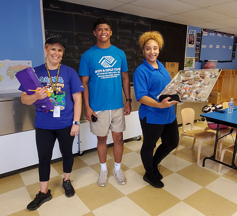 Three adults, all wearing blue shirts, stand and smile. The person on the right is holding a tray of food.