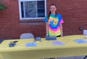 A woman wearing a tie dye tshirt with a PB on it smiles as she stands by a table.