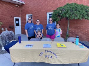Three adults and two children stand behind a table with a tan table cloth that says EJR Elementary PTA.