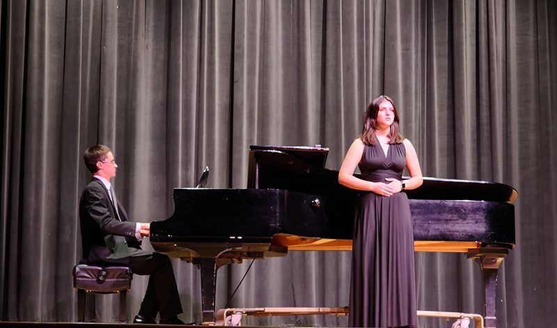 A young man dressed in a black suit sits at a large black piano playing while a young woman dressed in a long black gown, stands in front of it singing.