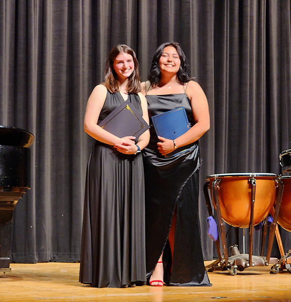 Two high school young women both dressed in long black gowns, standing on a stage.