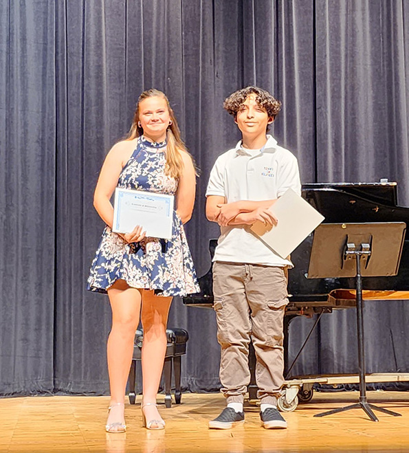 Two middle school students stand on a stage smiling. The girl on the left is wearing a blue print dress and has long blonde hair. The boy on the right has dark hair and is wearing a polo shirt and tan pants. They are both smiling and holding awards.