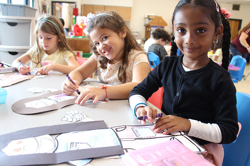 Three young elementary girls work at a table coloring with crayons.