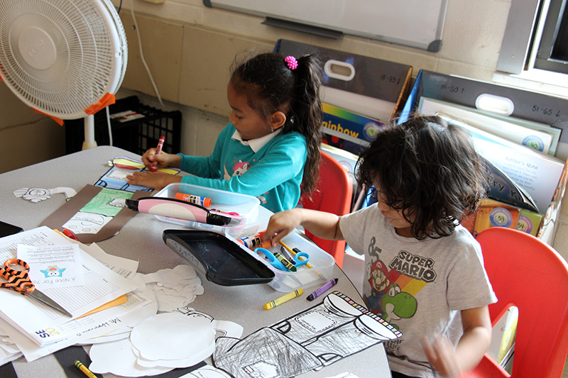 Two first grade students work on cutouts of themselves, coloring them and writing some of their favorite things.