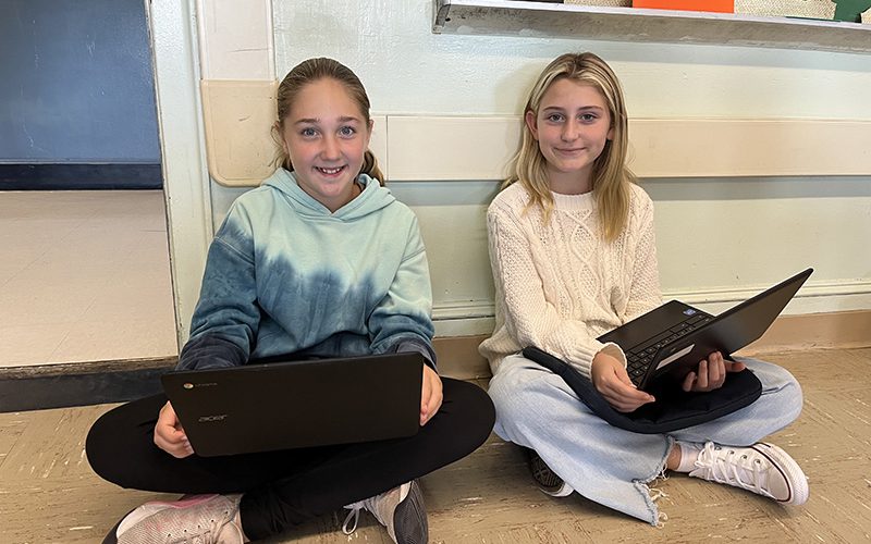 Two middle school age girls sit cross legged on the floor and smile. They are working on their Chromebooks