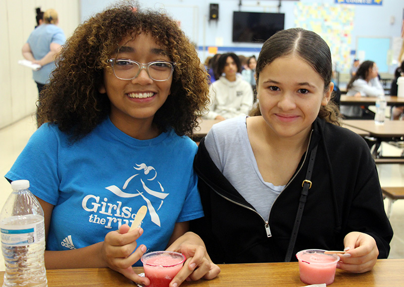Two seventh-grade girls sit at a table and smile as they eat red Italian ice. The girl on the left has long curly dark hair and is wearing glasses. The girl on the right has long dar hair pulled back in a ponytail and is wearing a light blue shirt and black zipped jacket.
