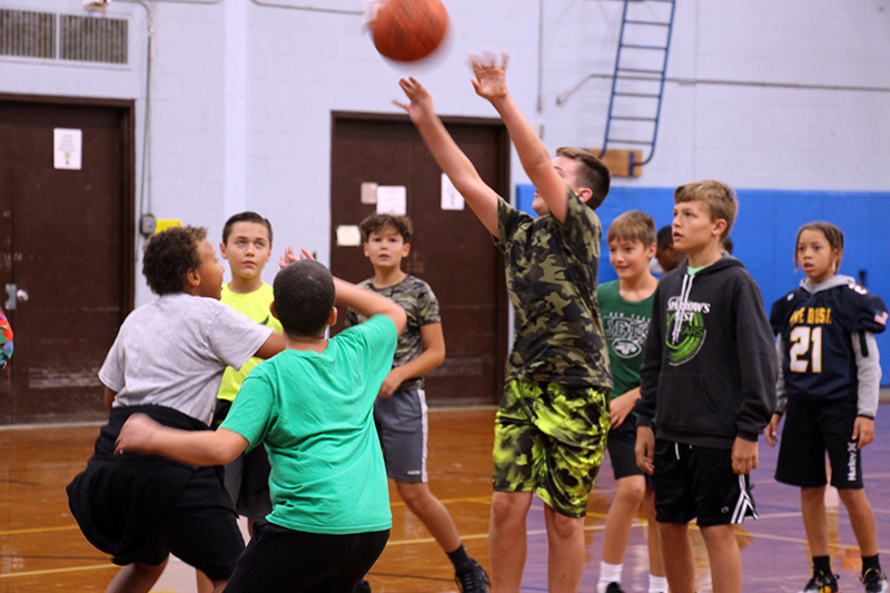 A group of sixth grade boys, most dressed in green, play basketball. One boy is shooting the ball into the air.