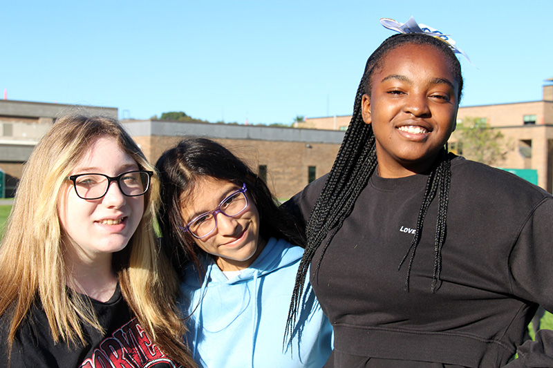 Three eighth-grade girls stand outside on a bright sunny day. they are smiling.