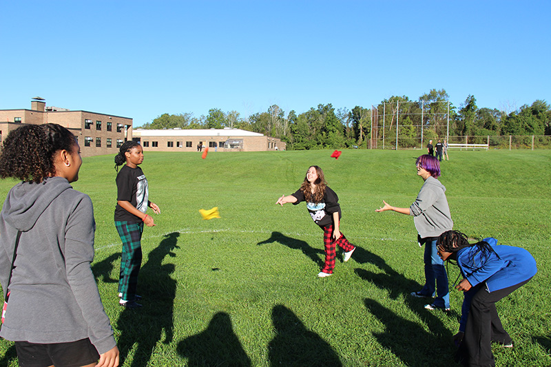 A group of five eighth-grade girls stand in a circle outside on a bright sunny day. They are tossing little discs to each other.